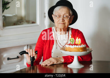 Respektable gut in Schwarz mini-hut grau behaarte ältere Frau mit Zuckerrohr ihren 90. Geburtstag bei Tisch zu Hause ist feiern gekleidet. Stockfoto