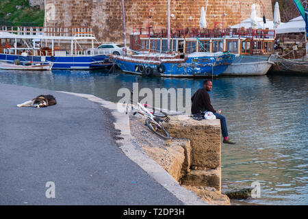 Kyrenia, Zypern - November 27, 2018: Ein afrikanischer Mann sitzt am Meer am alten hafen von Paphos auf Zypern ein Hund in der Nähe von ihn ist Schlafen auf der Ta Stockfoto