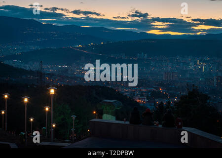 Drei junge Leute sitzen oben auf dem Hügel mit Blick auf die Hauptstadt von Georgien, Tiflis, während die Sonne hinter dem Hügel geht Stockfoto