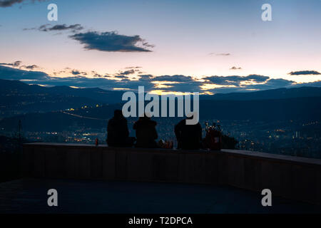 Drei junge Leute sitzen oben auf dem Hügel mit Blick auf die Hauptstadt von Georgien, Tiflis, während die Sonne hinter dem Hügel geht Stockfoto