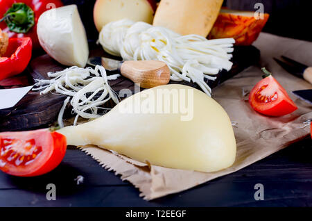 Sortierte hausgemachten Käse in verschiedenen Formen und Größen auf dem hölzernen Hintergrund, suluguni, Zopf, Cacocavallo mit Tomaten und Kräutern Stockfoto