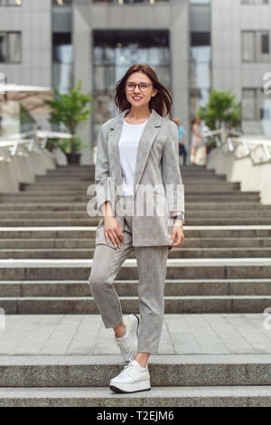 Junge Geschäftsfrau tragen Brillen stehen auf der Straße in die Kamera lächelt glücklich windiges Wetter full body shot Posing Stockfoto