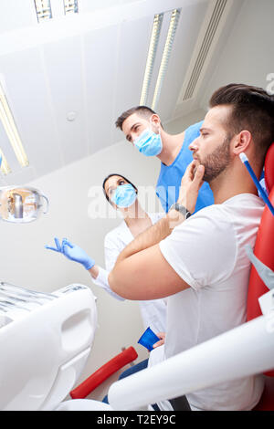 Porträt von einem gutaussehenden jungen Patienten in den Schmerz mit dem Zahnarzt und seinem Assistenten. Stockfoto