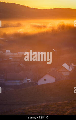 Schöne Szene mit ländlichen Häuser am Morgen mit farbiger Nebel von der Sunrise gegen trockene Bäume auf einem Hügel Stockfoto