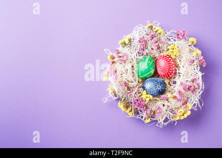 Von Hand bemalt, verschiedene Farben Ostereier im Nest und Blumen auf Violett Papier Hintergrund Stockfoto