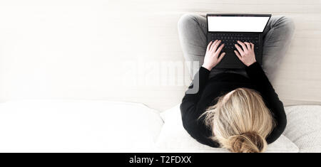 Direkt oberhalb der Schuß des blonden Jungen kaukasischen Frau mit gekreuzten Beinen sitzen auf einem Sofa Arbeiten am Laptop. Stockfoto