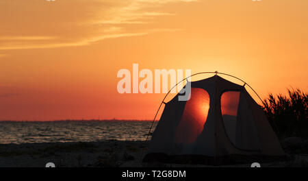 Panoramablick auf den schönen roten Sonnenuntergang am Meer und Camping Zelt mit glänzenden durch Eingang am Strand im Sommer Abend Stockfoto