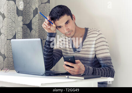 Portrait des jungen Mannes in Freizeitkleidung auf dem Job Arbeiten am Laptop, Smartphone und mit nachdenklichen Gesicht Stockfoto