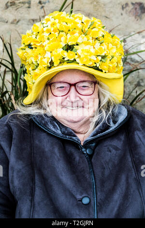 Eine ältere Dame lächelnd und tragen eine Neuheit Hut in der gelben Narzissen bedeckt. Stockfoto