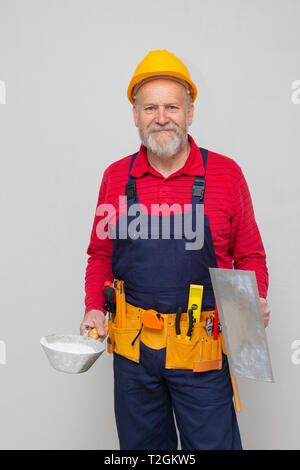 Ältere Konstruktor mit gelben Helm Holding Malwerkzeuge, Posieren vor der Beton Wand - neues Haus in Bau Stockfoto