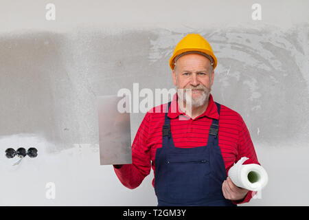 Ältere Konstruktor mit gelben Helm Holding Malwerkzeuge, Posieren vor der Beton Wand - neues Haus in Bau Stockfoto