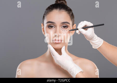 Plastische Chirurgie Konzept. Arzt Zeichnung Markierungen auf weibliche Gesicht, bevor Verfahren, grau studio Hintergrund Stockfoto