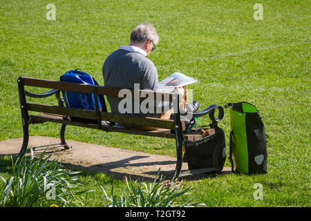 Ein Mann im Rentenalter sitzt in der Sonne auf einer Parkbank Lesen der Zeitung The Times in Hinksey Park, Oxford, UK Stockfoto