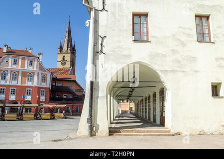 Alte, Mittelalterliche, weißes Gebäude mit einem Korridor mit mehreren Bögen und ein Kirchturm im Hintergrund, auf "Piata Mica" Platz - Sibiu (Hermannstadt Stockfoto