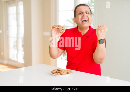 Mittleres Alter Mann essen Chocolate Chips cookies zu Hause schreien stolz und feiern Sieg und Erfolg sehr aufgeregt, jubelnde Emotion Stockfoto
