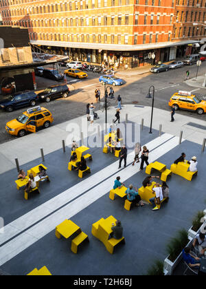Bei dieser Hochwinkelaufnahme werden Menschen in einem öffentlichen Außenbereich um gelbe Tische sitzen und stehen gesehen. Die Szene spielt in einer Stadt mit Gebäuden und Straßen im Hintergrund. Die Leute genießen wahrscheinlich den öffentlichen Raum, vielleicht essen, plaudern oder einfach nur entspannen. Die gelben Tabellen verleihen der Szene einen Farbakzent und machen sie optisch ansprechend. Stockfoto