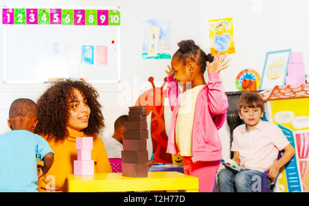 Kleine schwarze Mädchen, lockiges Haar spielen Bausteine mit Lehrer im Kindergarten Klasse heben die Hände, glücklich Stockfoto