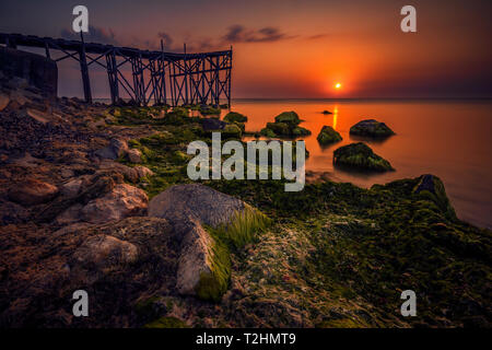 Sonnenaufgang vom Schwarzen Meer in Rumänien gesehen mit langen Exposition mit Steinen in Algen im Vordergrund verdeckt und eine hölzerne Brücke Ponton in der backgro Stockfoto