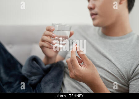 Ein junger Mann ist krank mit Grippe, liegt zu Hause unter einer Decke, nimmt eine Pille Stockfoto
