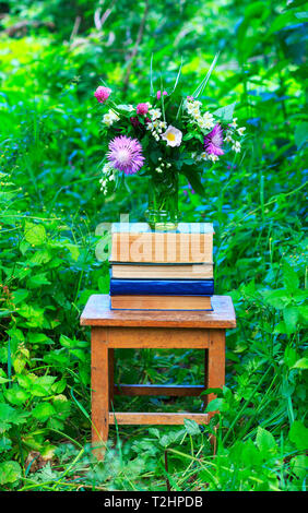 Ein Blumenstrauß von Clover, Kornblumen und Jasmin in einem Glas Vase und einen Stapel Bücher auf einem alten Holzstuhl unter dem grünen Gras in der autolöscher Stockfoto