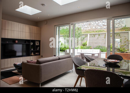 Ein modernes Doppelzimmer und Wohnraum in der Rückseite eines Hauses mit einer Couch, großem Fernseher und Esstisch mit Veranda und Garten. Stockfoto