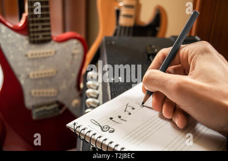 Junger Mann Komponieren von Musik in einem Personal Notebook mit Gitarre und Bass im Hintergrund. Musiker schreiben im Music Studio. Komponieren im Violinschlüssel. Stockfoto