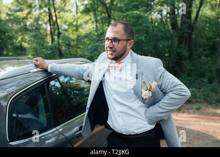 Der Bräutigam mit Bart in einer grauen Jacke und Brille steht in Anlehnung an das Auto. Stockfoto