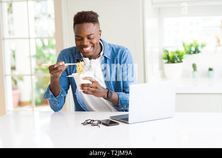 Hübscher junger African Business Mann essen Lieferung asiatische Lebensmittel und Arbeiten mit Computer, genießen Nudeln lächelnd Stockfoto