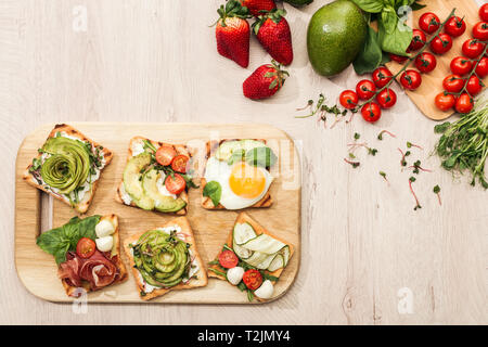 Blick von oben auf die Toasts mit Gemüse auf Schneidebrett und frische Zutaten auf hölzernen Tisch Stockfoto
