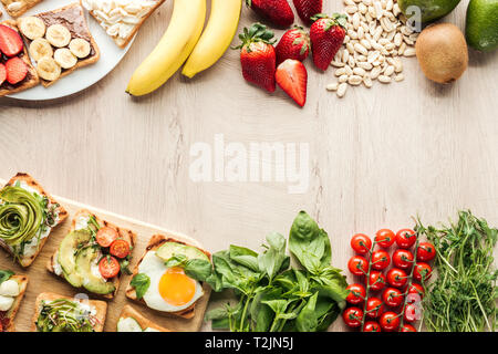 Blick von oben auf die Zutaten auf Holztisch und Toast mit Gemüse auf Schneidebrett Stockfoto