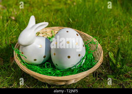 Ein Osterhase und Ostereier aus weißer Keramik mit goldenen Polka Dots in einem Korb auf einer Wiese. Stockfoto