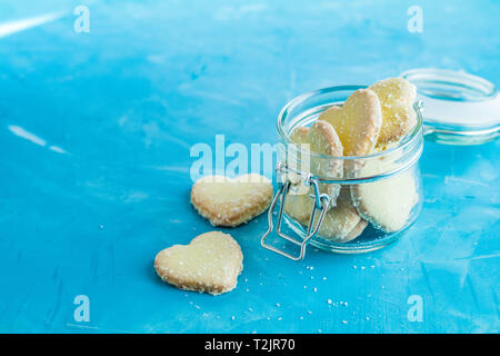 Köstliche gebackene Kekse in Herzform mit Kokos Chips in Glas Glas auf blauen Beton Tabelle Oberfläche. Essen Grußkarte mit kopieren Sie Text. Stockfoto
