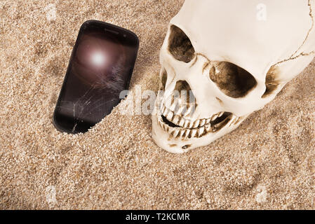 Nahaufnahme auf Skull über Sand Hintergrund. Gebrochene Smartphone an seiner Seite. Stockfoto