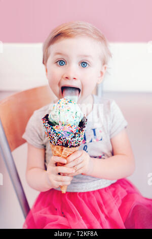 Lustige Kaukasischen blond babyl Mädchen mit blauen Augen Essen lecken Eis in große Waffel Kegel mit Streuseln und zeigt Zunge Süß. Happy chi Stockfoto