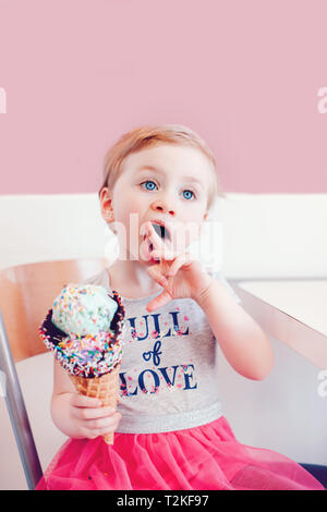 Cute adorable lustig Kaukasischen blond babyl Mädchen mit blauen Augen Eis essen in große Waffel Kegel mit bunten Streuseln. Kid lecken Finger Stockfoto