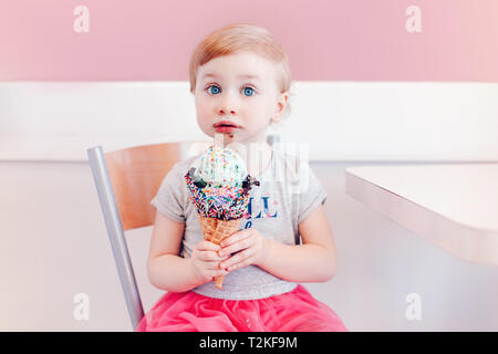 Cute adorable lustig Kaukasischen blond babyl Mädchen mit blauen Augen Essen lecken Eis in große Waffel Kegel mit bunten Streuseln. Happy Chil Stockfoto