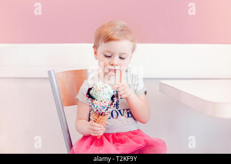 Cute adorable lustig Kaukasischen blond babyl Mädchen mit blauen Augen Essen lecken Eis in große Waffel Kegel mit bunten Streuseln. Happy Chil Stockfoto