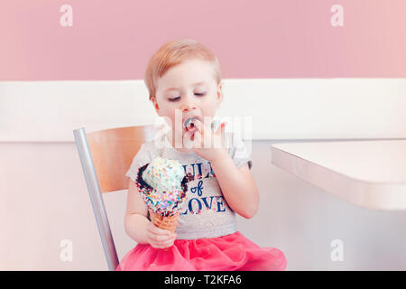 Cute adorable lustig Kaukasischen blond babyl Mädchen mit blauen Augen Eis essen in große Waffel Kegel mit bunten Streuseln. Kid lecken Finger Stockfoto