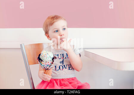 Cute adorable lustig Kaukasischen blond babyl Mädchen mit blauen Augen Eis essen in große Waffel Kegel mit bunten Streuseln. Kid lecken Finger Stockfoto