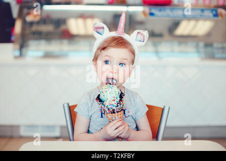 Cute adorable lustig Kaukasischen blonde Mädchen mit den blauen Augen tragen Einhorn stirnband Essen lecken Eis in große Waffel Kegel mit Streuseln. Stockfoto