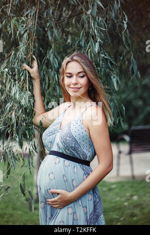 Glückliche gesunde Schwangerschaft und Mutterschaft. Portrait von Schwangeren jungen kaukasischen Frau mit langen blauen Kleid in Park außerhalb posieren. Schöne nachdenkliche Fe Stockfoto