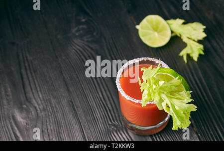 Cocktail Bloody Mary mit Kalk und Sellerie, Salz und Snacks in einem Glas auf einem schwarzen Holztisch. Stockfoto