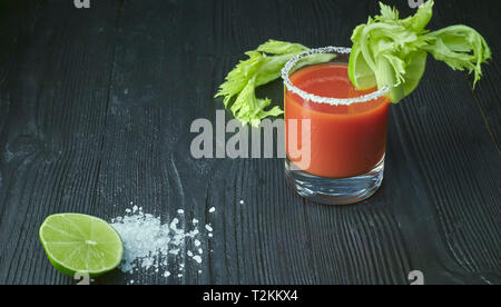 Cocktail Bloody Mary mit Kalk und Sellerie, Salz und Snacks in einem Glas auf einem schwarzen Holztisch. Stockfoto