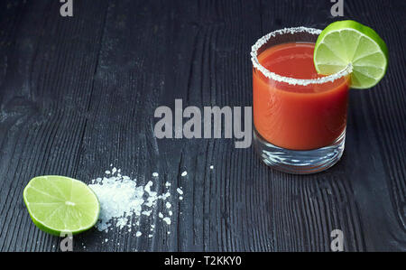 Cocktail Bloody Mary mit Kalk und Sellerie, Salz und Snacks in einem Glas auf einem schwarzen Holztisch. Stockfoto