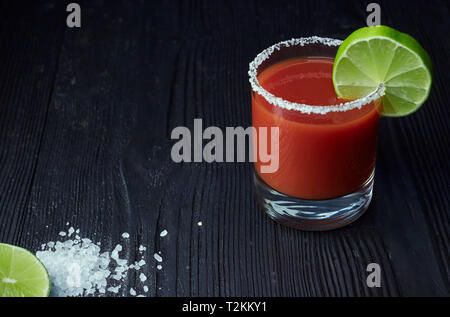 Cocktail Bloody Mary mit Kalk und Sellerie, Salz und Snacks in einem Glas auf einem schwarzen Holztisch. Stockfoto