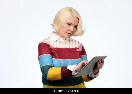 Gerne ältere blonde Frau mit Verwendung der digitalen Tablet surfen Web social media online chatten. Europäische Dame, Internet Shopping auf Weiß Grau st Stockfoto