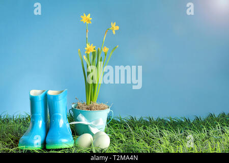 Natürliche Ostereier, Gummi regen Stiefeln und gelben Narzisse Blumen im Gras vor einem blauen Hintergrund mit Raum für Kopieren. Stockfoto