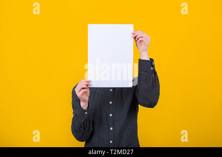 Geschäftsfrau, die ihr Gesicht mit einem leeren weißen Blatt Papier, wie eine Maske für das Verstecken ihrer Identität über gelb Wand Hintergrund isoliert. Introvertiert f Stockfoto