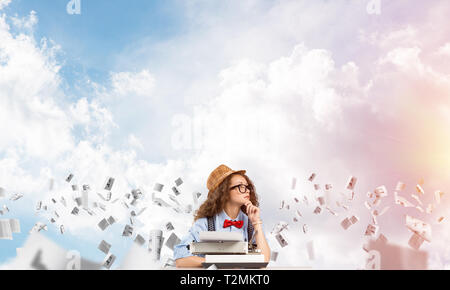 Junge und schöne Frau Schriftsteller in Hut und Brille mit Eingabe von Maschine, während am Tisch unter fliegende Papiere mit bewölkt skyscape auf bak-Sitzung Stockfoto