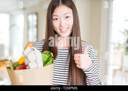 Schöne asiatische Frau mit papiertüte von frischen Lebensmitteln zufrieden mit Lächeln tun ok Zeichen, Daumen hoch, mit den Fingern, ausgezeichnete Zeichen Stockfoto